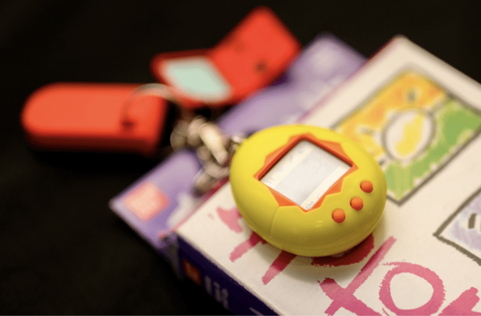 Yellow Tamagotchi virtual pet on its box, with a red flip-phone keychain behind it